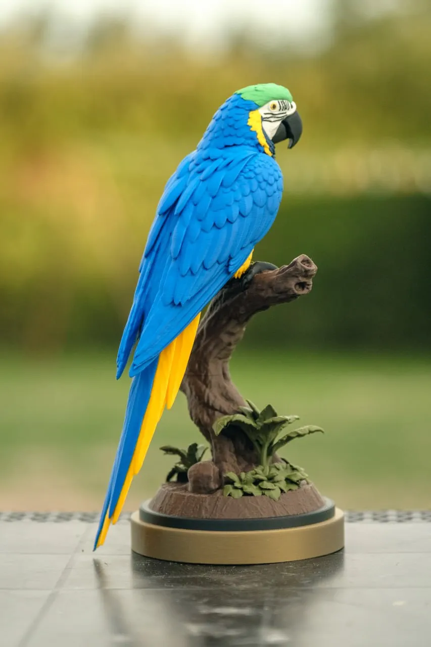 Blue-and-yellow parrot statue perched on a branch with foliage and a round base